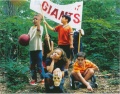 a scan of the John Henry CD tray artwork. Depicted are 6 kids together surrounded by bushes and trees. Two of them stand, holding up old up a large white banner that reads 'THEY MIGHT BE GIANTS' in red capital letters. A girl hunches forward behind them, holding the red seat of a toy wagon. In front of those three are two more kids: a boy poking at the ground sitting on the detached wheels and axle of the same toy wagon, and another girl crouched down, resting her head on her left hand and holding a human skull on top of her knee with her right. The word ‘THEY’ is written on the skull’s forehead in black. Finally, to their left, another boy stands, holding a red rubber ball in one arm and raising the other in the air.